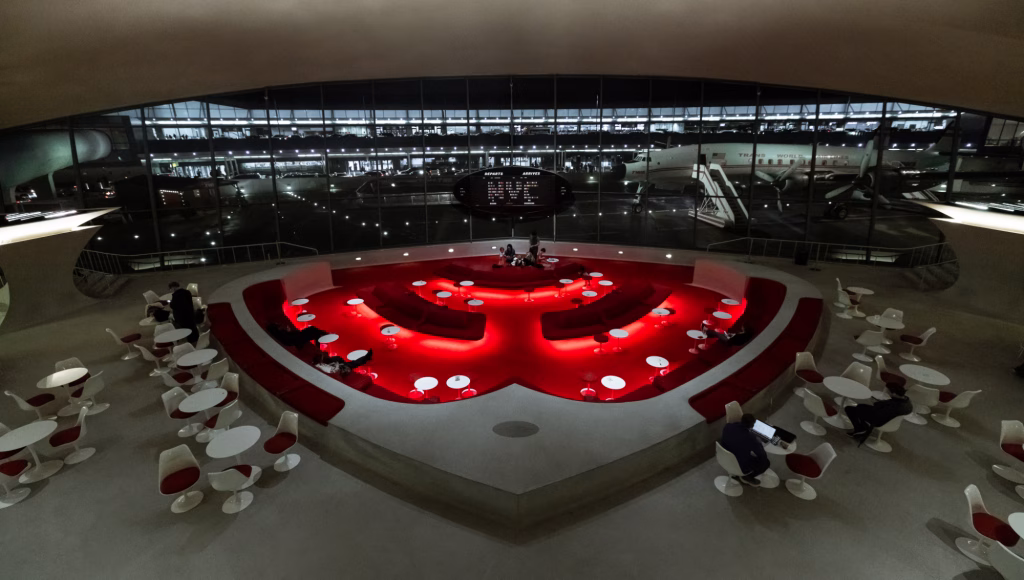 Interior of the TWA Hotel at JFK with a red sunken lounge, white tables, and expansive airfield views, highlighting custom millwork in the restored mid-century terminal.